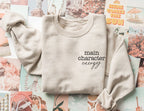folded sand-colored sweatshirt that says main character energy, styled on pastel photo collage