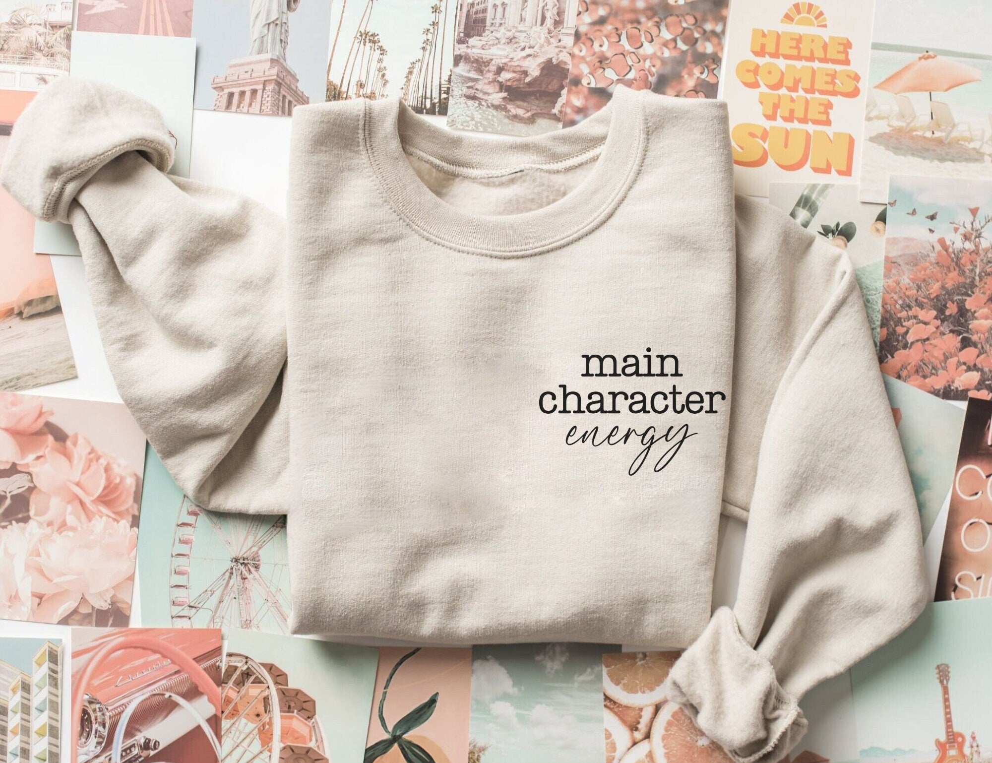 folded sand-colored sweatshirt that says main character energy, styled on pastel photo collage