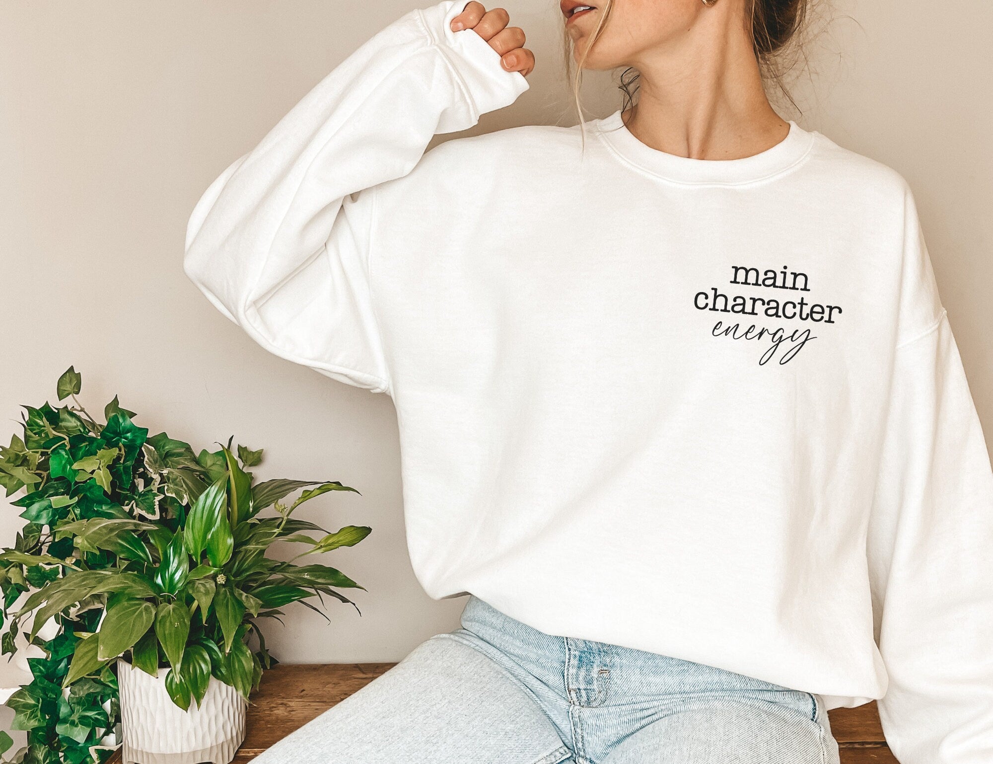 woman wearing white sweatshirt that says main character energy sitting beside green indoor plant