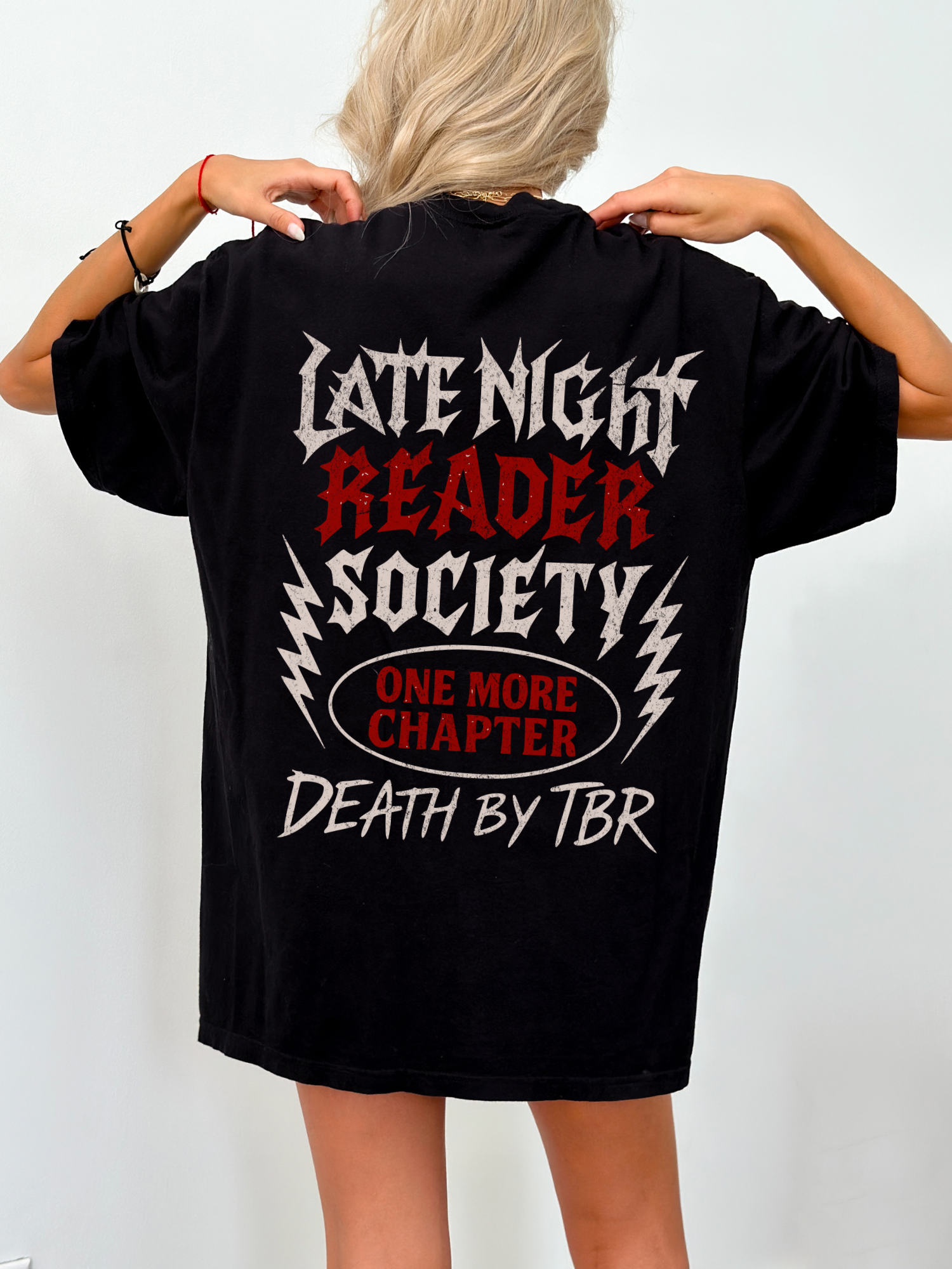 Oversized black t-shirt worn by a blonde woman, showcasing a bold metal-inspired back design with the text “Late Night Reader Society,” “One More Chapter,” and “Death by TBR” in grungy gothic fonts. Ideal gift for fantasy bookworms who stay up reading.