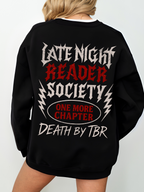 A black sweatshirt with a bold heavy metal-inspired graphic that says Late Night Reader Society in distressed white and red letters, surrounded by lightning bolts. Centered below is a red oval with the words One More Chapter and underneath it reads Death by TBR. The sweatshirt is worn oversized on a blonde model standing with her back to the camera.