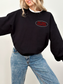 A closeup of the front of the black sweatshirt worn by a smiling woman. The front features a small red oval with the words One More Chapter printed on the left chest. She wears blue jeans and a white tee underneath, with her arms lifted in a playful pose.