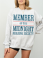 woman wearing an ash grey sweatshirt with large blue and red text that says member of the midnight reading society