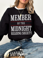 woman sitting cross-legged in a black sweatshirt that says member of the midnight reading society in white and red text
