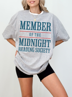 woman wearing a athletic heather grey t-shirt that says member of the midnight reading society in teal blue and red graphic text