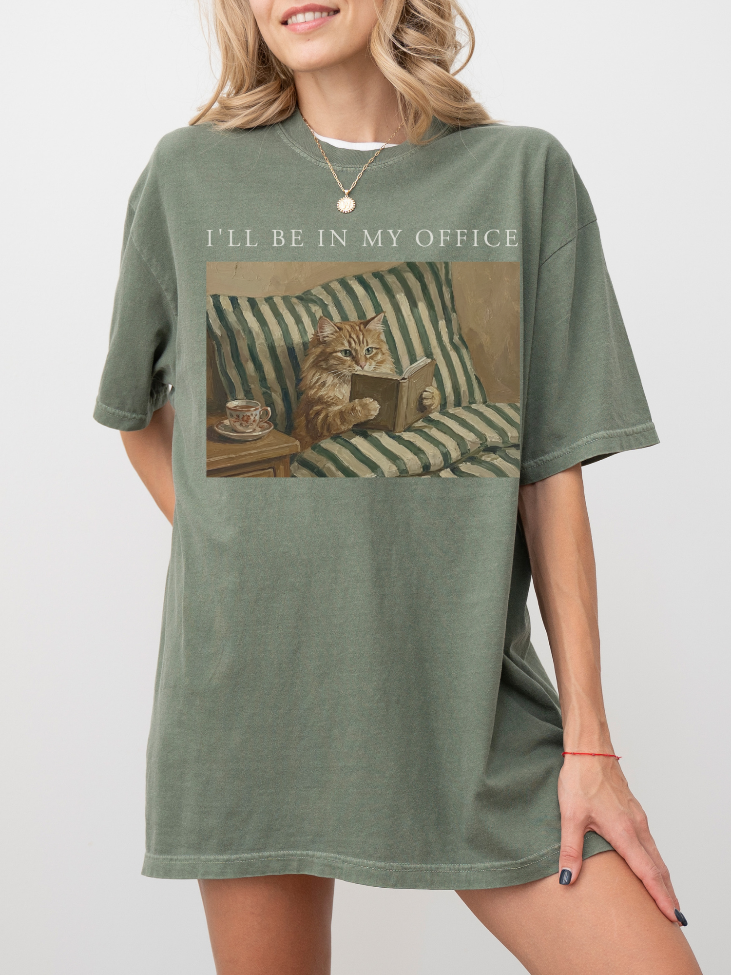 Moss oversized t shirt with Ill be in my office text and a cozy cat reading in bed illustration. Styled on a model with a relaxed fit creating a soft casual outfit for readers, introverts, and cat lovers.
