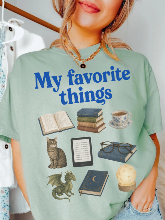 Woman wearing a bay Comfort Colors shirt featuring My Favorite Things design with books, coffee, cat, Kindle, dragon, glasses, moon journal, and cozy reading essentials for book lovers