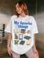 Woman wearing a white Comfort Colors shirt featuring My Favorite Things design with books, coffee, cat, Kindle, dragon, glasses, moon journal, and cozy reading essentials for readers