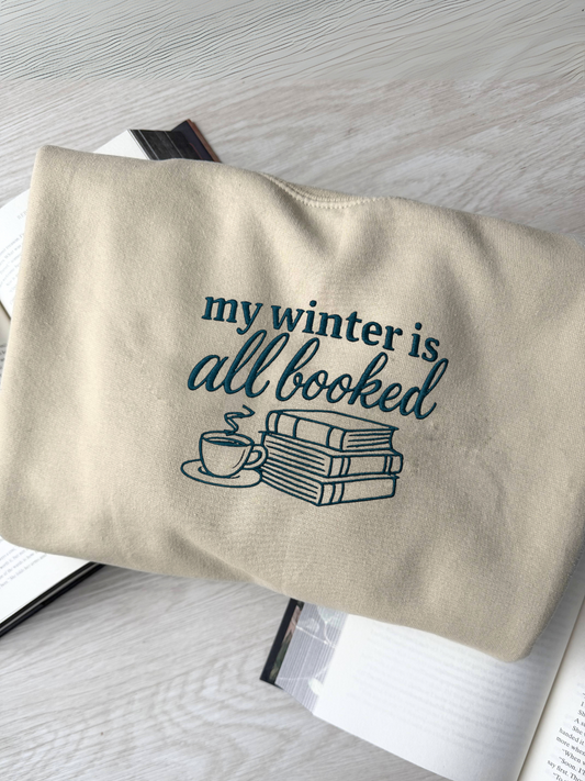 Embroidered sand color sweatshirt with "my winter is all booked" design featuring books and a coffee cup, laid on an open book background for cozy winter reading vibes

