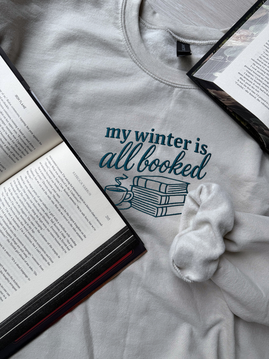 Flatlay of embroidered white color sweatshirt with "my winter is all booked" design surrounded by books and a white scrunchie for cozy bookworm aesthetic

