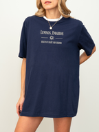 Model wearing a navy blue t-shirt with cream print that reads "Lumnos, Emarion - Realm of Light and Shadow", styled with a gold pendant and white underlayer, standing against a white backdrop

