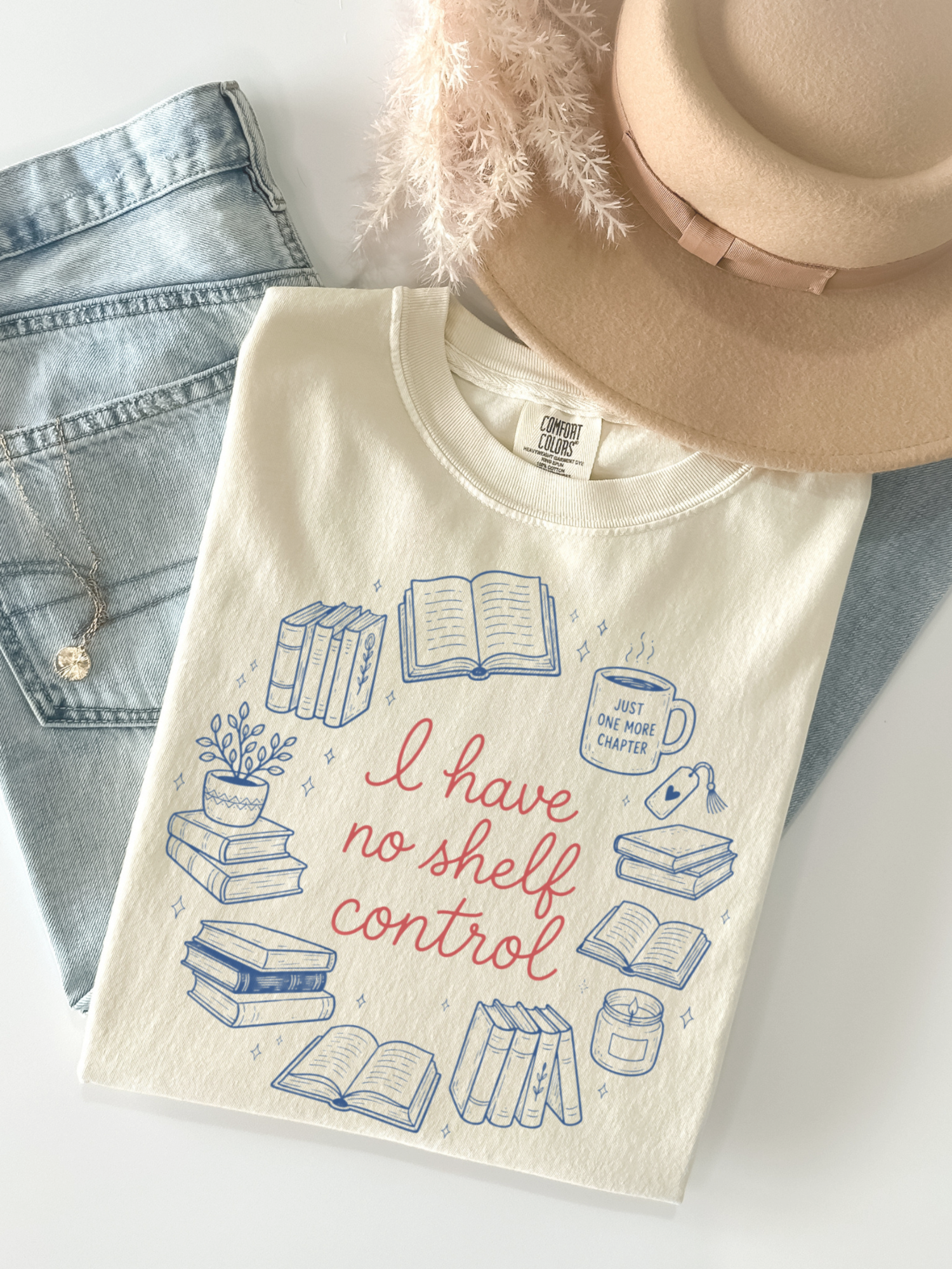 Flat lay of ivory Comfort Colors t shirt with blue book illustrations and red text I have no shelf control surrounded by stacks of books coffee mug and reading icons styled with denim jeans and beige hat