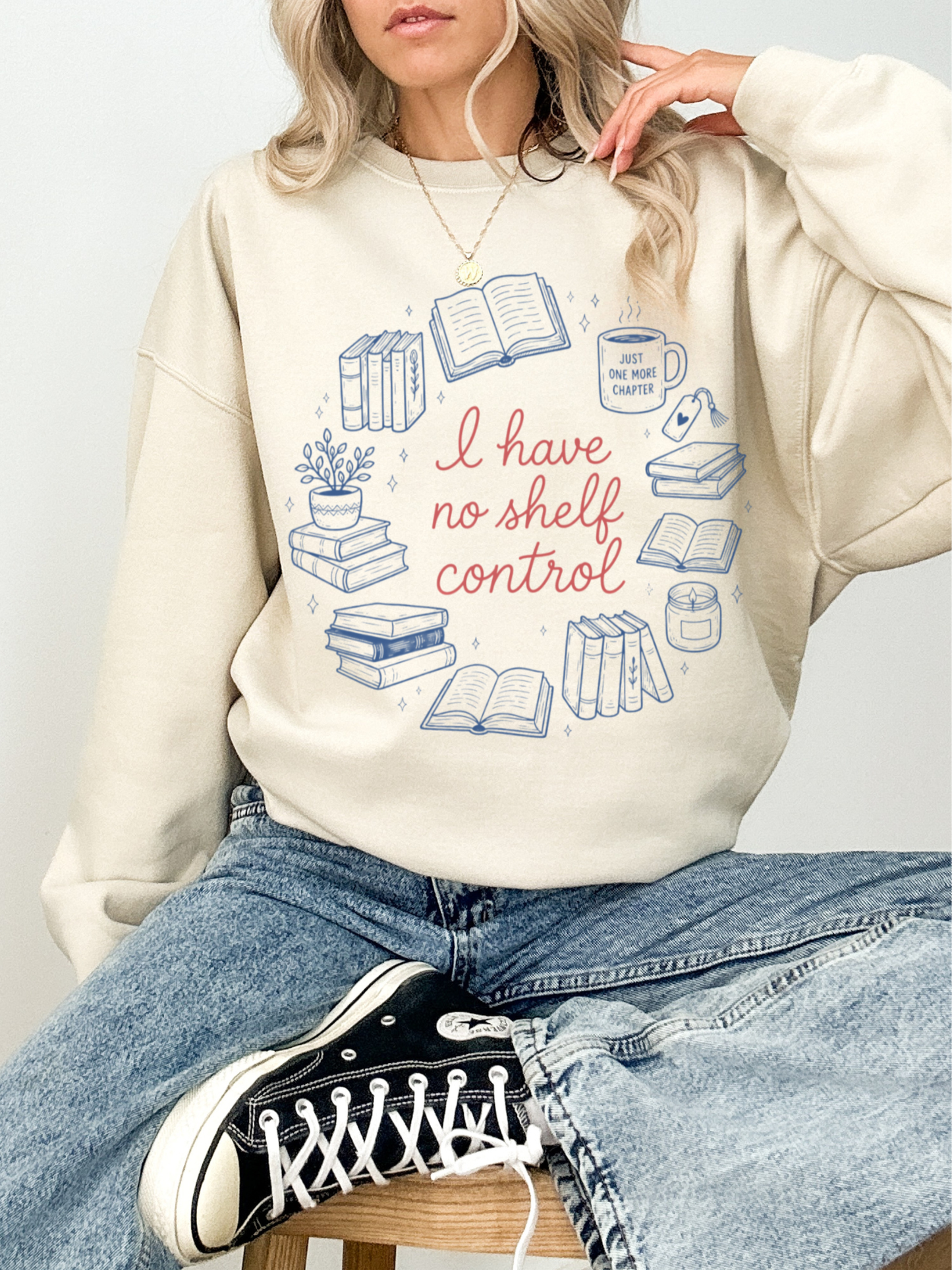Woman sitting on stool wearing sand colored sweatshirt with I have no shelf control design surrounded by book illustrations styled with jeans and black sneakers