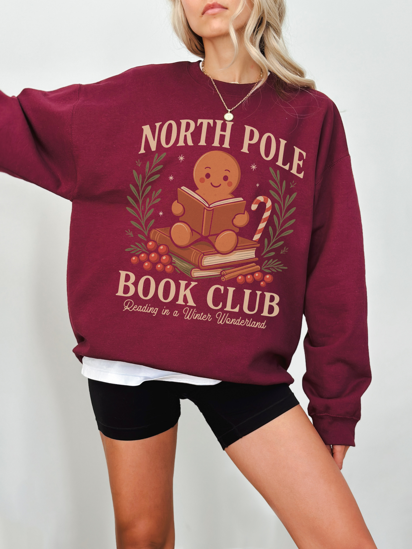 Woman wearing maroon North Pole Book Club sweatshirt featuring a cute gingerbread character reading a book surrounded by holly berries and a candy cane