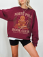 Woman wearing maroon North Pole Book Club sweatshirt featuring a cute gingerbread character reading a book surrounded by holly berries and a candy cane