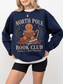Model wearing navy blue North Pole Book Club sweatshirt with gingerbread design, books, and Christmas accents