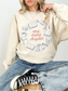 Woman sitting casually wearing sand sweatshirt with one more chapter design featuring book illustrations styled with relaxed jeans and sneakers