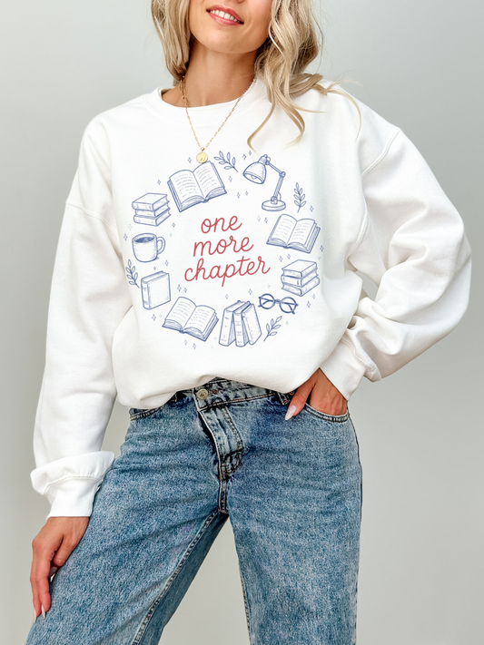 Woman wearing white sweatshirt with one more chapter text and blue book themed illustrations including books lamp and coffee mug styled with high waisted jeans