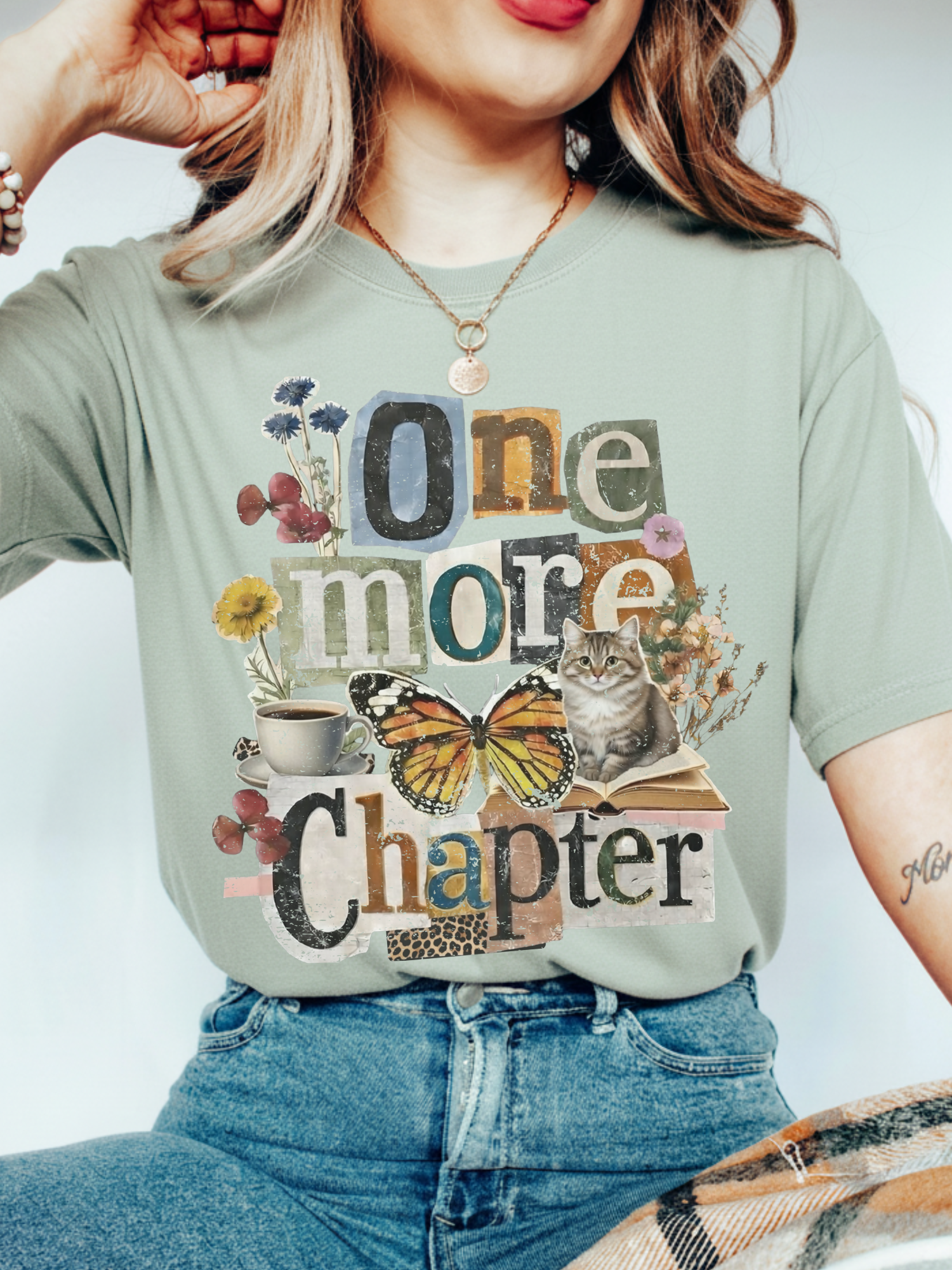 A woman wearing a bay green graphic t-shirt with a collage design that reads 'One more Chapter' featuring a butterfly, cat, flowers, and coffee cup, paired with blue jeans.