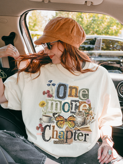 A woman sitting in a car wearing an ivory graphic t-shirt with 'One More Chapter' written in a retro collage font with butterflies and flowers, paired with a brown corduroy cap and sunglasses.