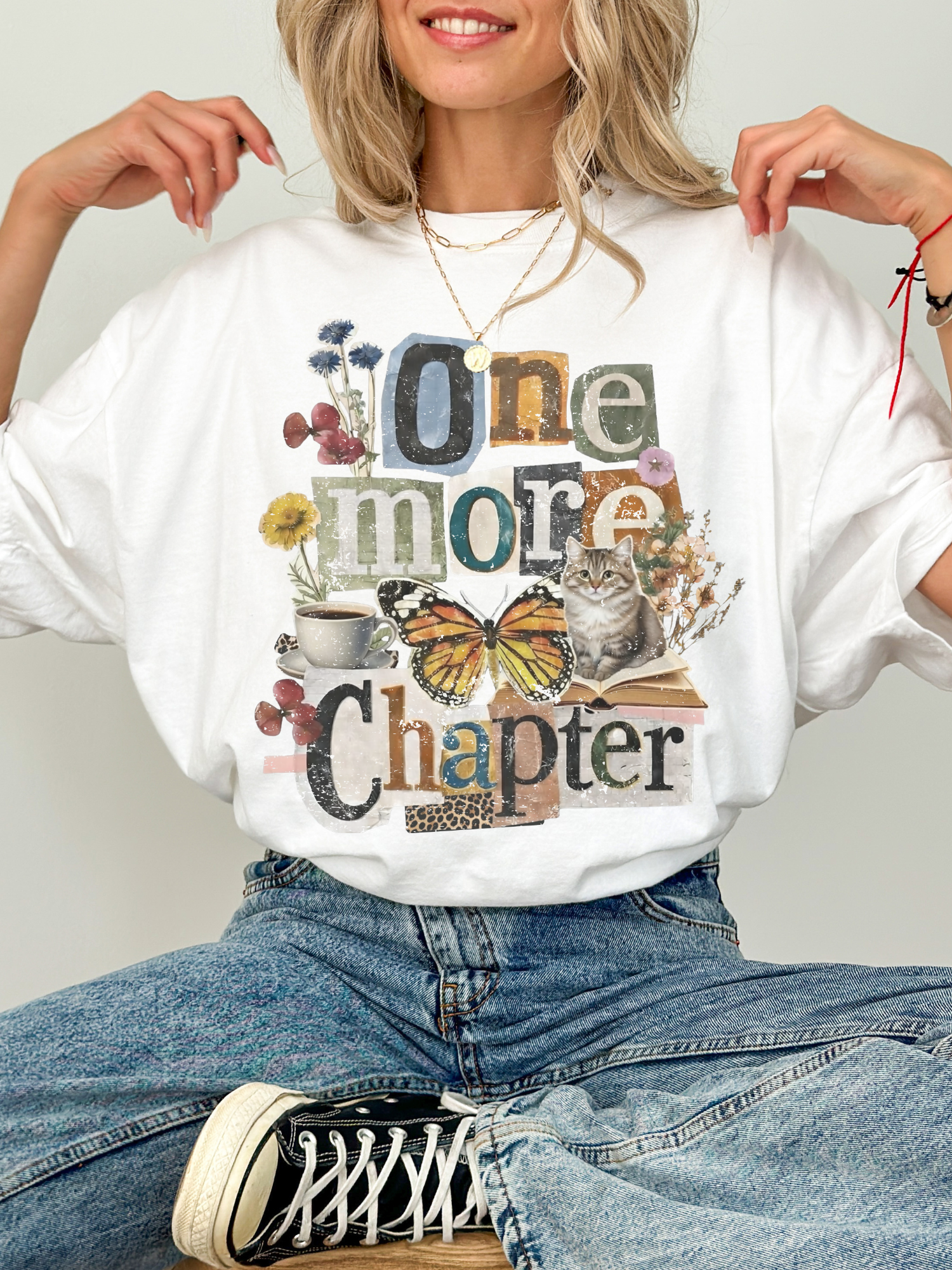 A woman wearing a white graphic t-shirt with a vintage-style collage print that reads 'One more Chapter' with a butterfly, cat, flowers, and coffee cup. She is wearing blue jeans and a black and white sneaker, sitting against a light gray background.