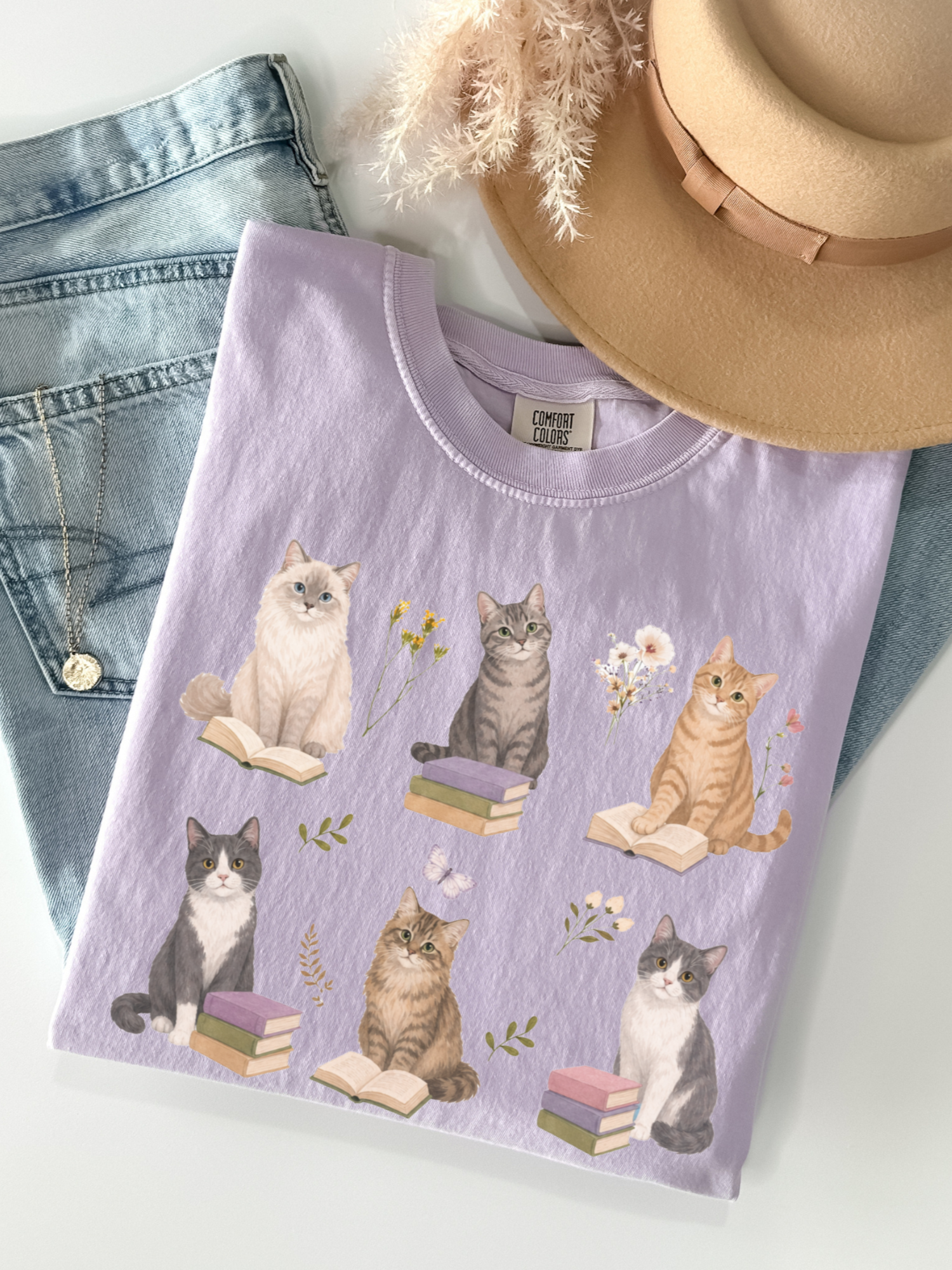 Flat lay of orchid Comfort Colors t shirt with illustrated cats sitting on books surrounded by flowers styled with denim jeans necklace and beige hat