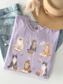 Flat lay of orchid Comfort Colors t shirt with illustrated cats sitting on books surrounded by flowers styled with denim jeans necklace and beige hat