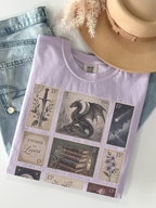 Flat lay of orchid Comfort Colors fantasy themed t shirt with vintage postage stamp collage featuring dragon moon antique books flowers and enemies to lovers design with jeans and hat