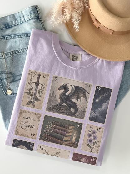 Flat lay of orchid Comfort Colors fantasy themed t shirt with vintage postage stamp collage featuring dragon moon antique books flowers and enemies to lovers design with jeans and hat
