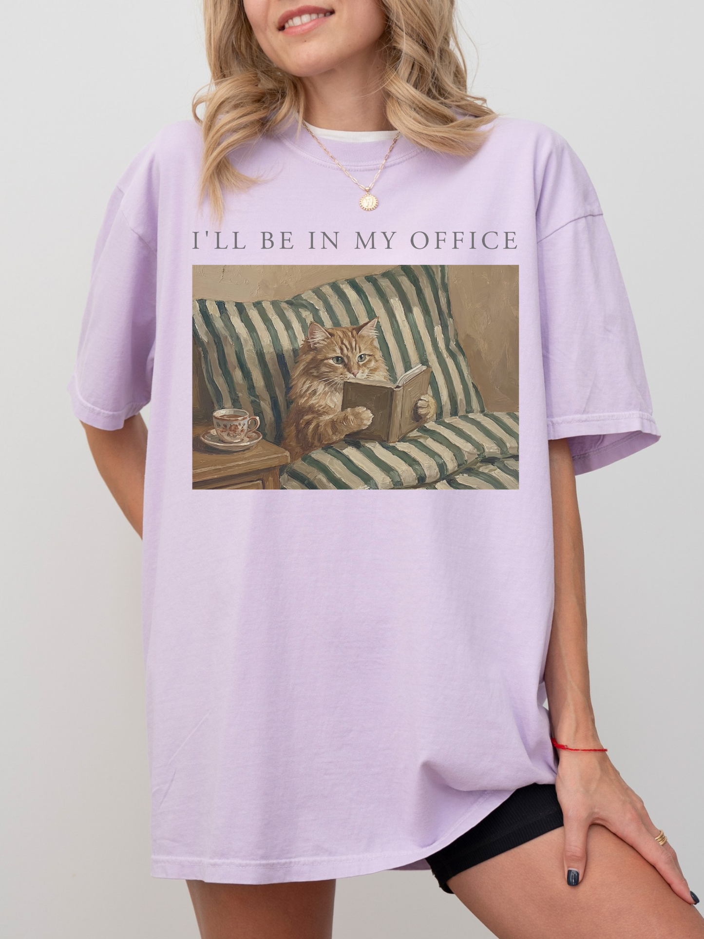 Orchid oversized t shirt with the phrase Ill be in my office and a cozy cat reading a book illustration with striped bedding and tea cup detail. Soft aesthetic outfit for readers, introverts, and cat lovers who enjoy book themed apparel.