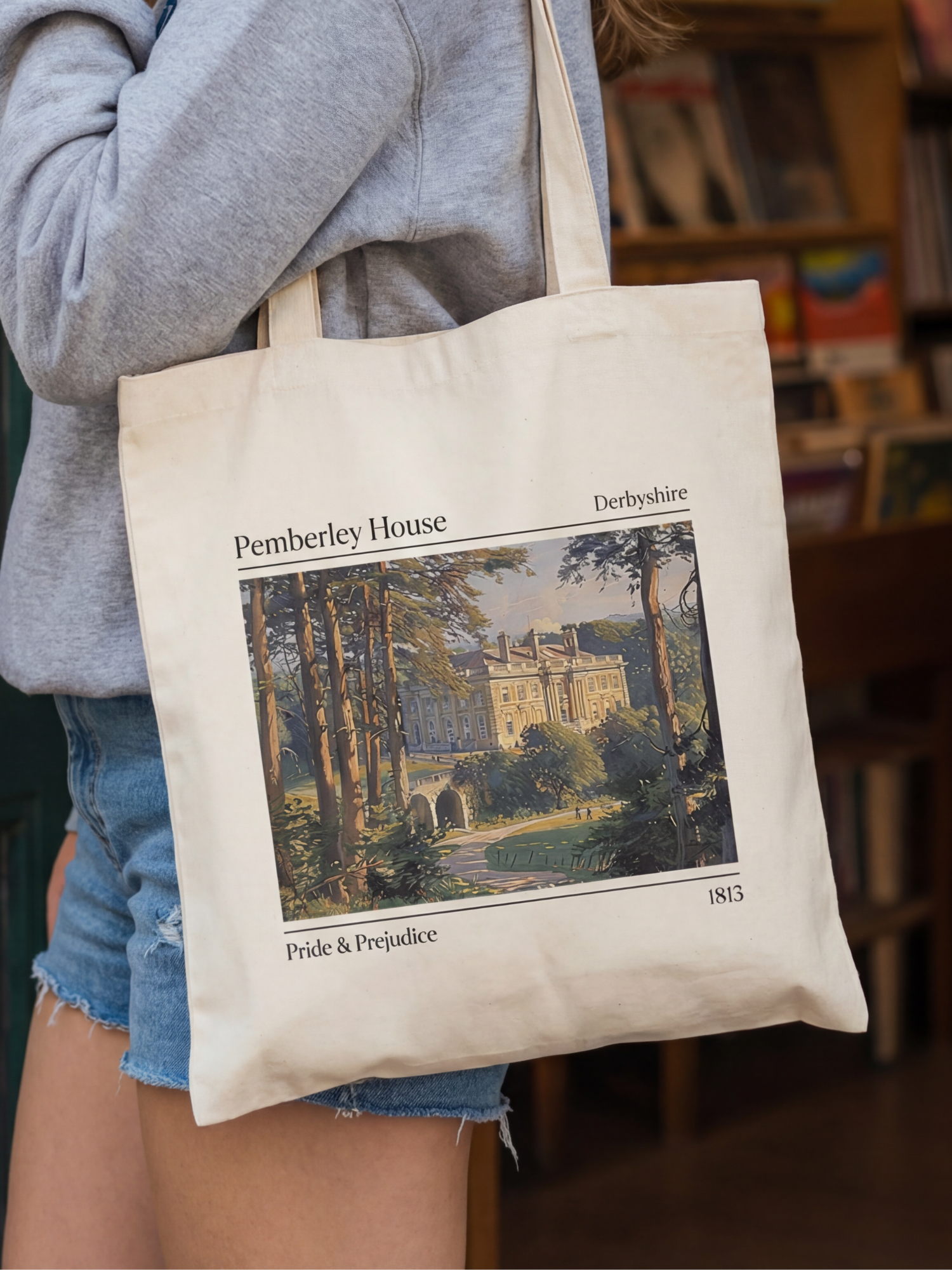 Canvas tote bag featuring a Pemberley House illustration inspired by Pride and Prejudice, showing a classic estate with trees and text details. The tote is worn in a cozy bookstore setting with shelves in the background, creating a literary aesthetic perfect for classic book lovers, readers, and fans of romantic novels.