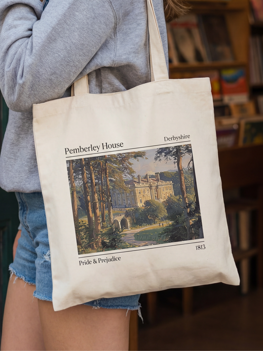 Canvas tote bag featuring a Pemberley House illustration inspired by Pride and Prejudice, showing a classic estate with trees and text details. The tote is worn in a cozy bookstore setting with shelves in the background, creating a literary aesthetic perfect for classic book lovers, readers, and fans of romantic novels.