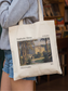 Canvas tote bag featuring a Pemberley House illustration inspired by Pride and Prejudice, showing a classic estate with trees and text details. The tote is worn in a cozy bookstore setting with shelves in the background, creating a literary aesthetic perfect for classic book lovers, readers, and fans of romantic novels.