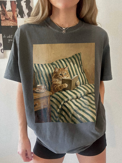 Pepper t-shirt featuring a detailed painting style cat reading a book while sitting in bed with striped blankets and a tea cup. Styled in a relaxed indoor setting with a cozy and slightly moody aesthetic, ideal for book lovers and cat themed apparel fans.