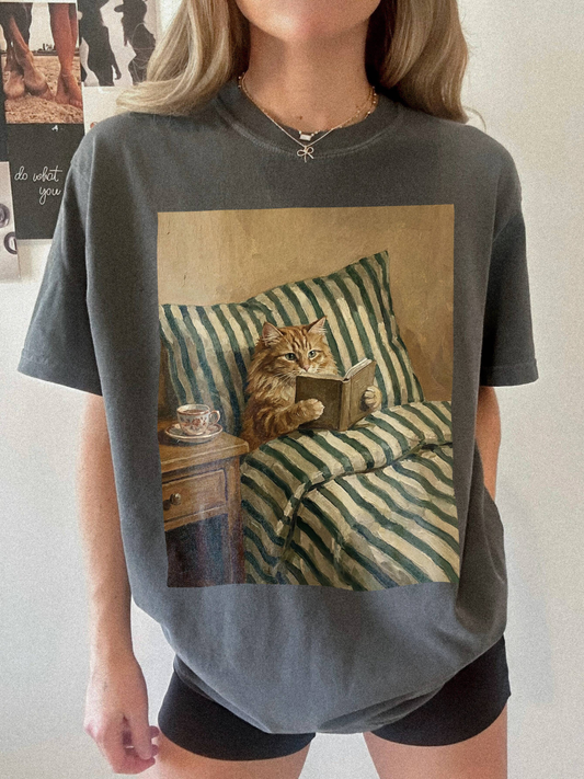 Pepper t-shirt featuring a detailed painting style cat reading a book while sitting in bed with striped blankets and a tea cup. Styled in a relaxed indoor setting with a cozy and slightly moody aesthetic, ideal for book lovers and cat themed apparel fans.