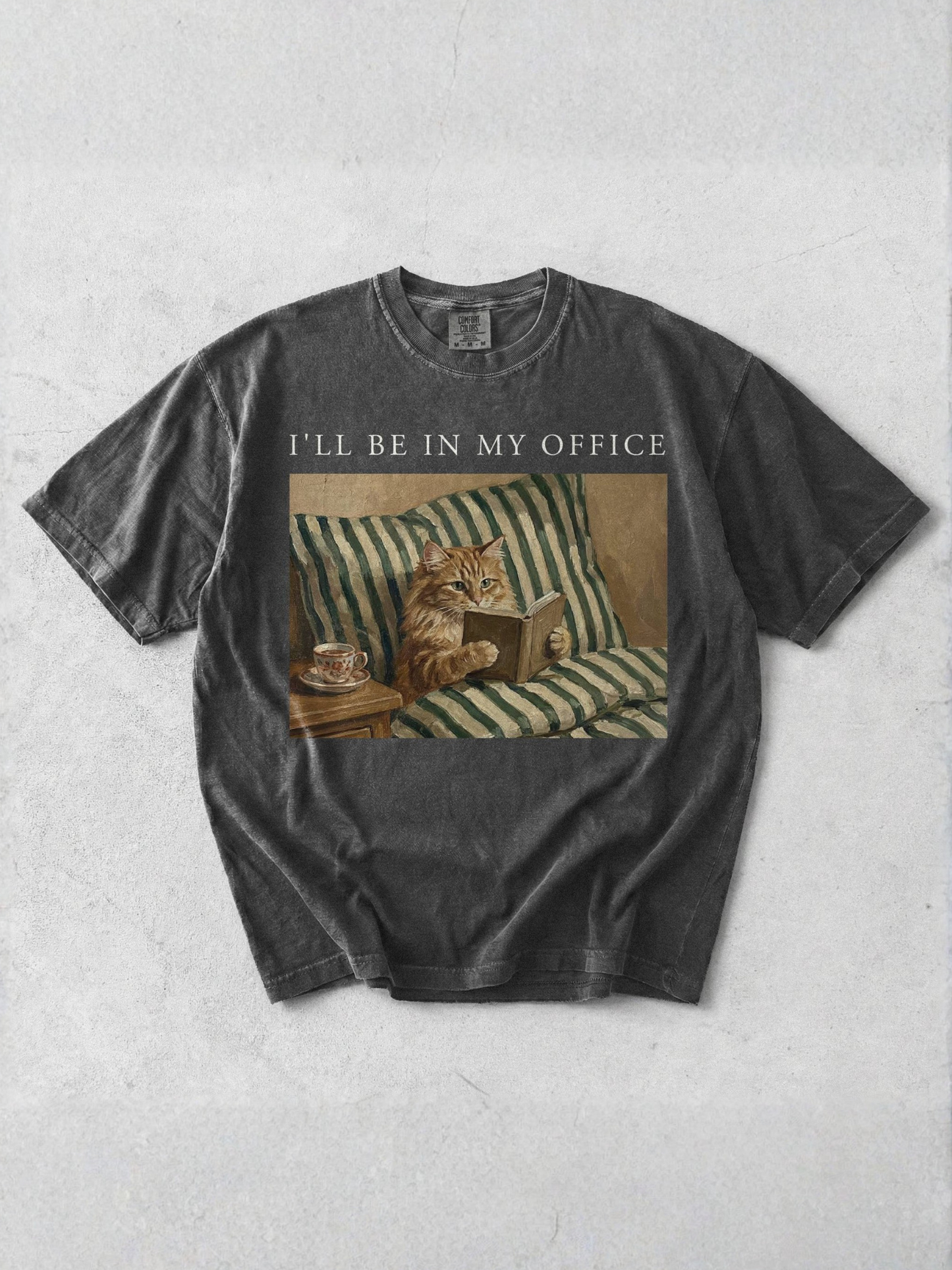 Pepper t shirt laid flat featuring Ill be in my office text and a detailed vintage style cat reading a book illustration. Minimal mockup highlighting a cozy reading theme shirt for book lovers and cat themed apparel.