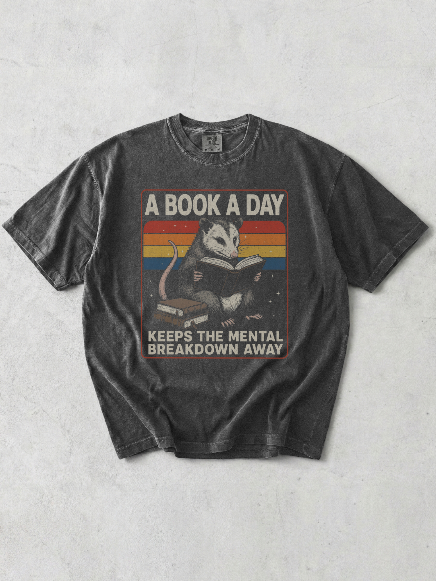 Flat lay of pepper Comfort Colors t shirt featuring opossum reading book with vintage rainbow stripes and book lover quote styled on neutral backdrop