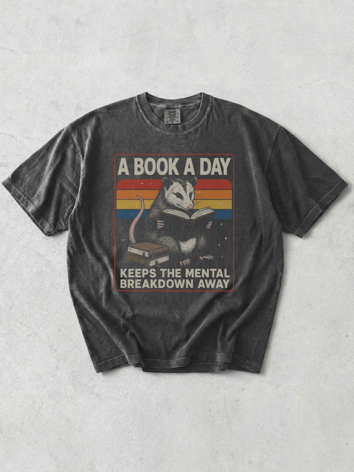 Flat lay of pepper Comfort Colors t shirt featuring opossum reading book with vintage rainbow stripes and book lover quote styled on neutral backdrop