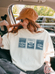 woman sitting in car wearing cream t shirt with Pride and Prejudice stamp illustration featuring Longbourn House Pemberley House and Netherfield Park casual outfit