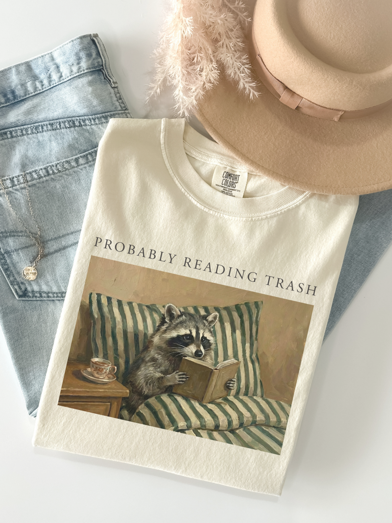folded ivory comfort colors shirt with probably reading trash raccoon reading book design styled with denim jeans beige hat and neutral aesthetic flatlay