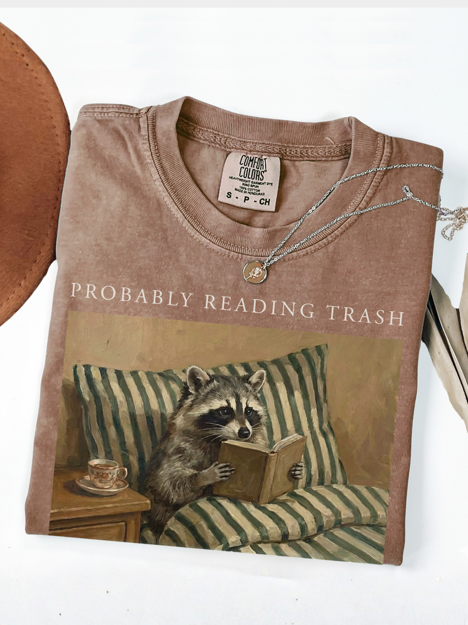 folded brown comfort colors shirt with probably reading trash raccoon reading book design styled with minimal neutral background cozy bookish aesthetic