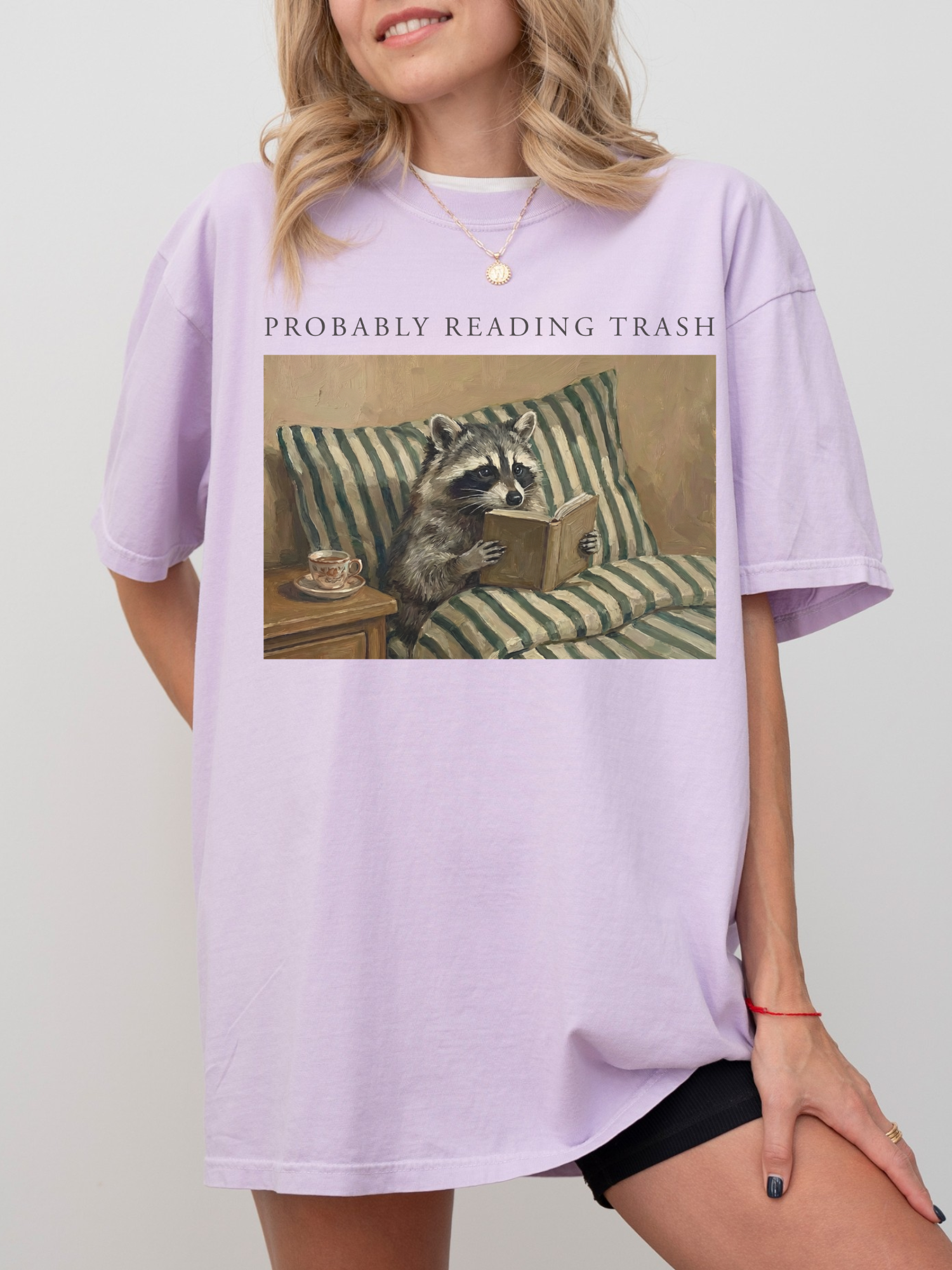 woman wearing oversized lavender shirt with probably reading trash text and vintage raccoon reading a book on a striped couch cozy aesthetic book lover apparel