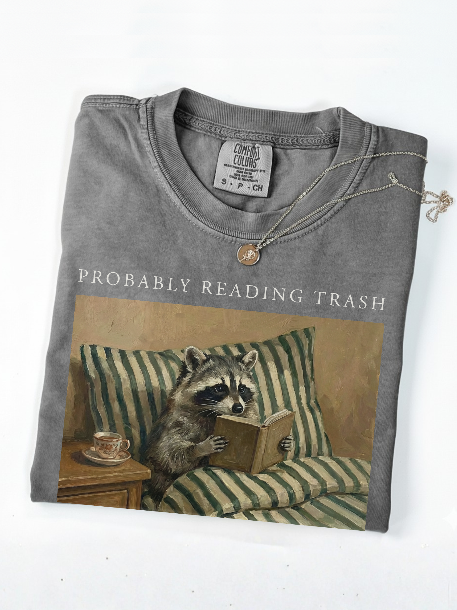 folded grey comfort colors shirt featuring probably reading trash raccoon reading book graphic with necklace detail close up product image
