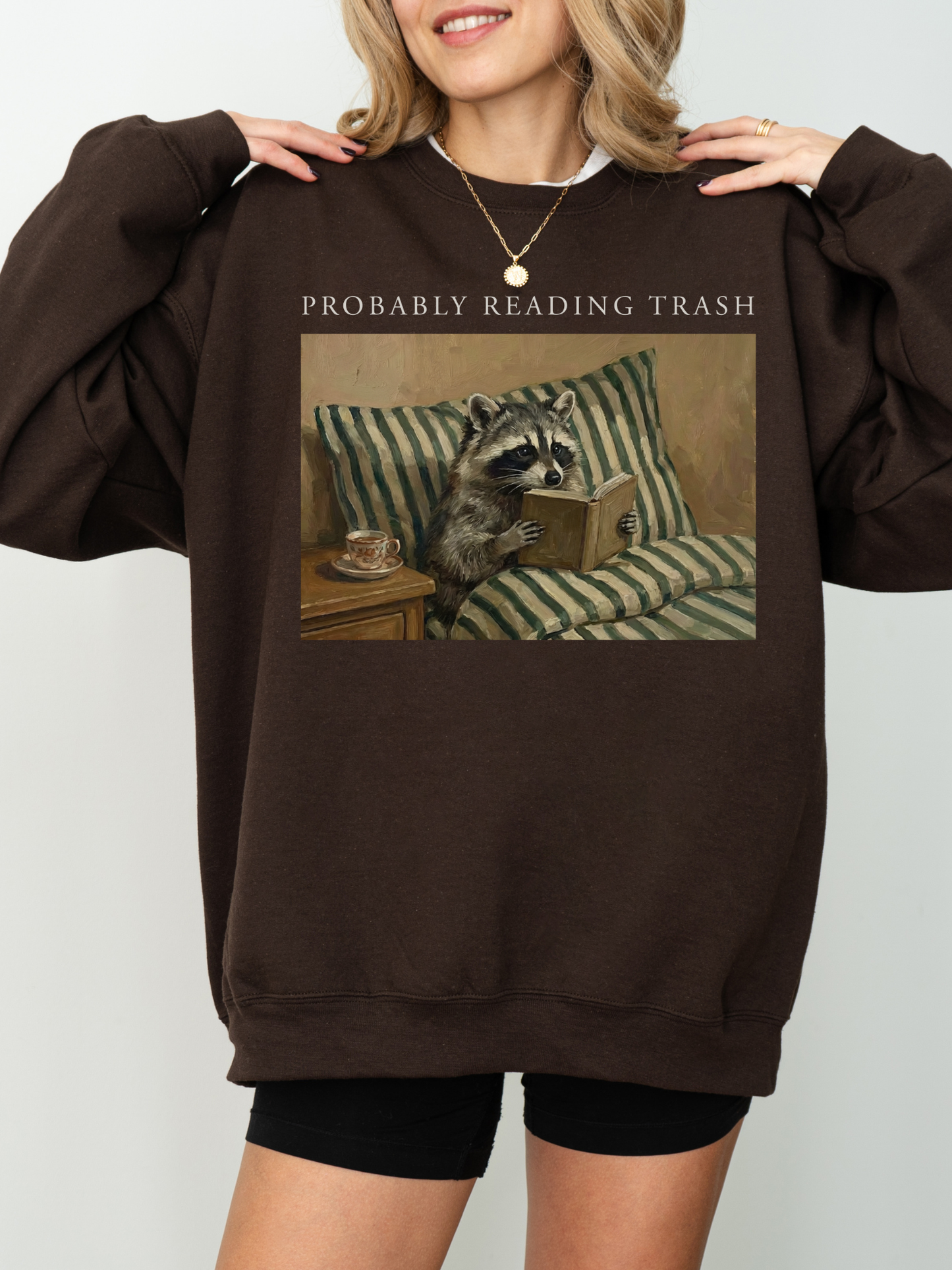 woman wearing oversized brown sweatshirt with probably reading trash raccoon reading book design warm cozy aesthetic book lover apparel