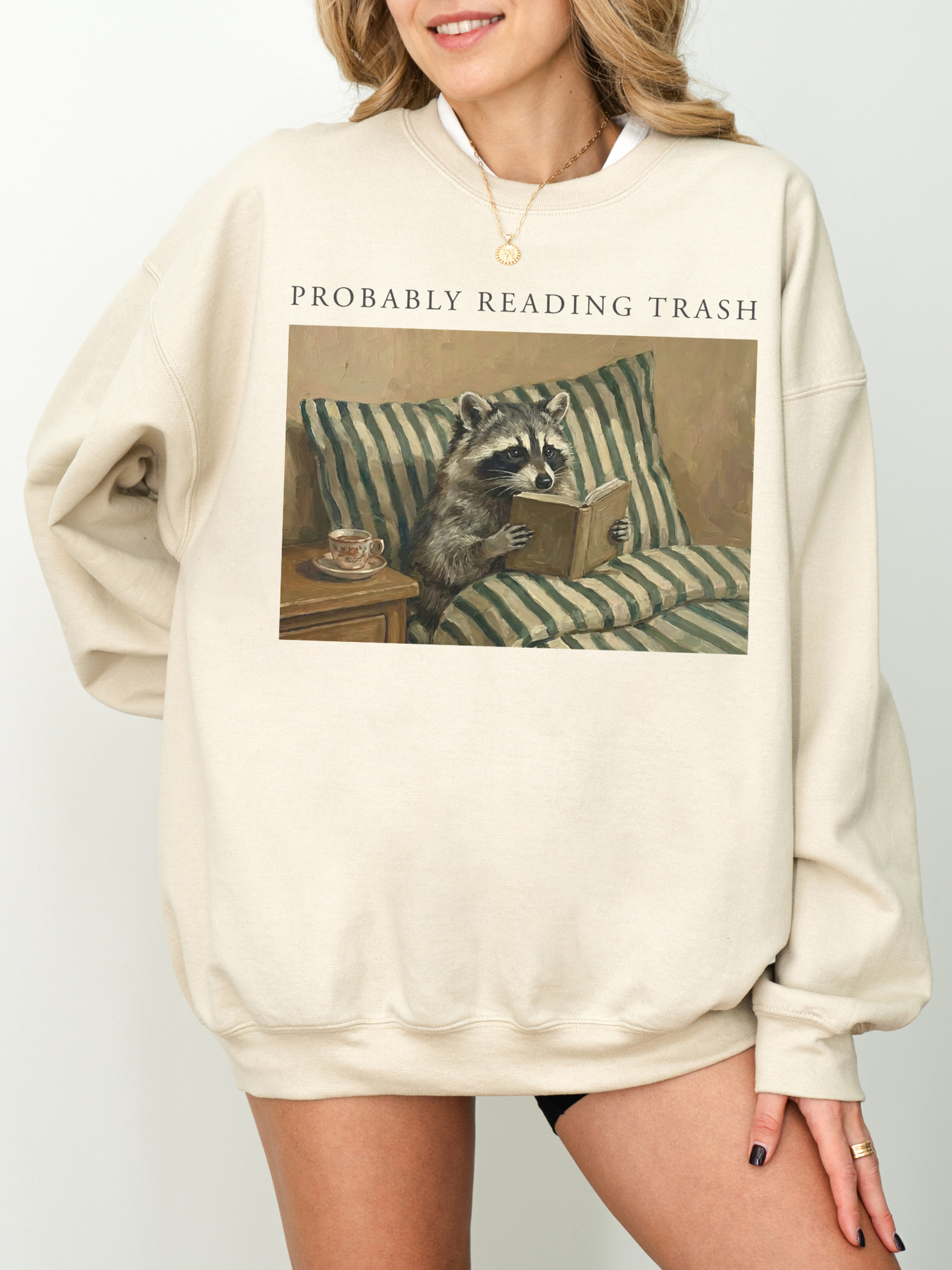 woman wearing oversized sand sweatshirt with probably reading trash text and vintage raccoon reading a book on striped couch cozy book lover aesthetic