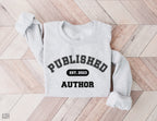 folded ash grey sweatshirt that says published author est 2023 on light wood floor
