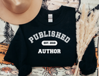 folded black sweatshirt that says published author est 2019 styled with beige hat and dried grass