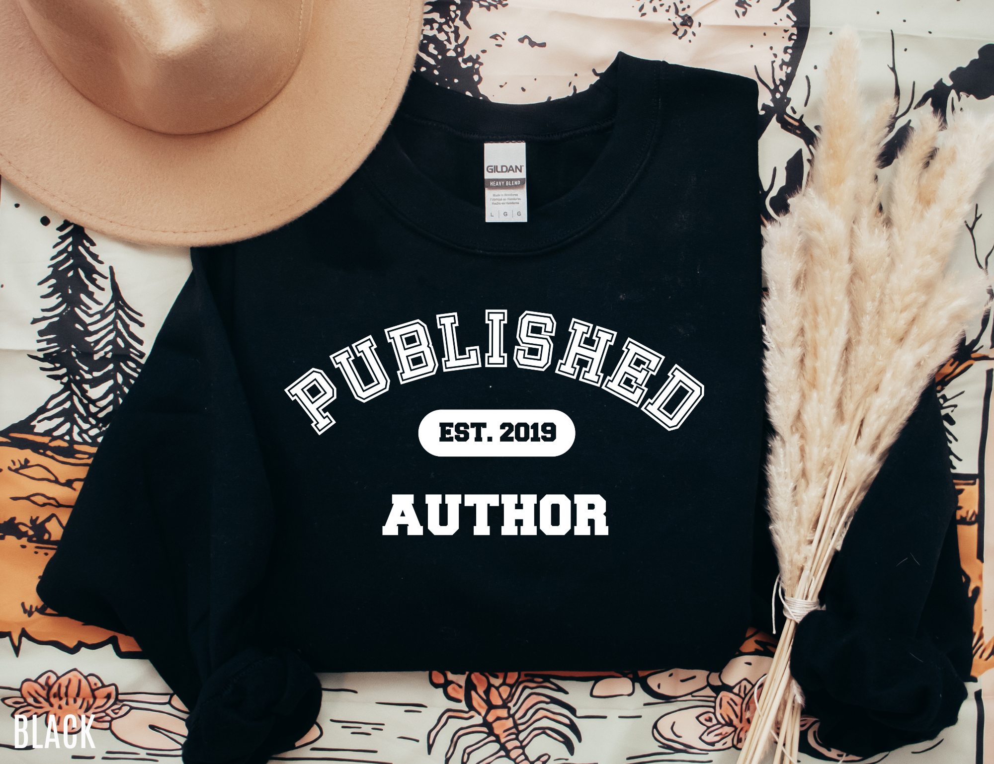 folded black sweatshirt that says published author est 2019 styled with beige hat and dried grass