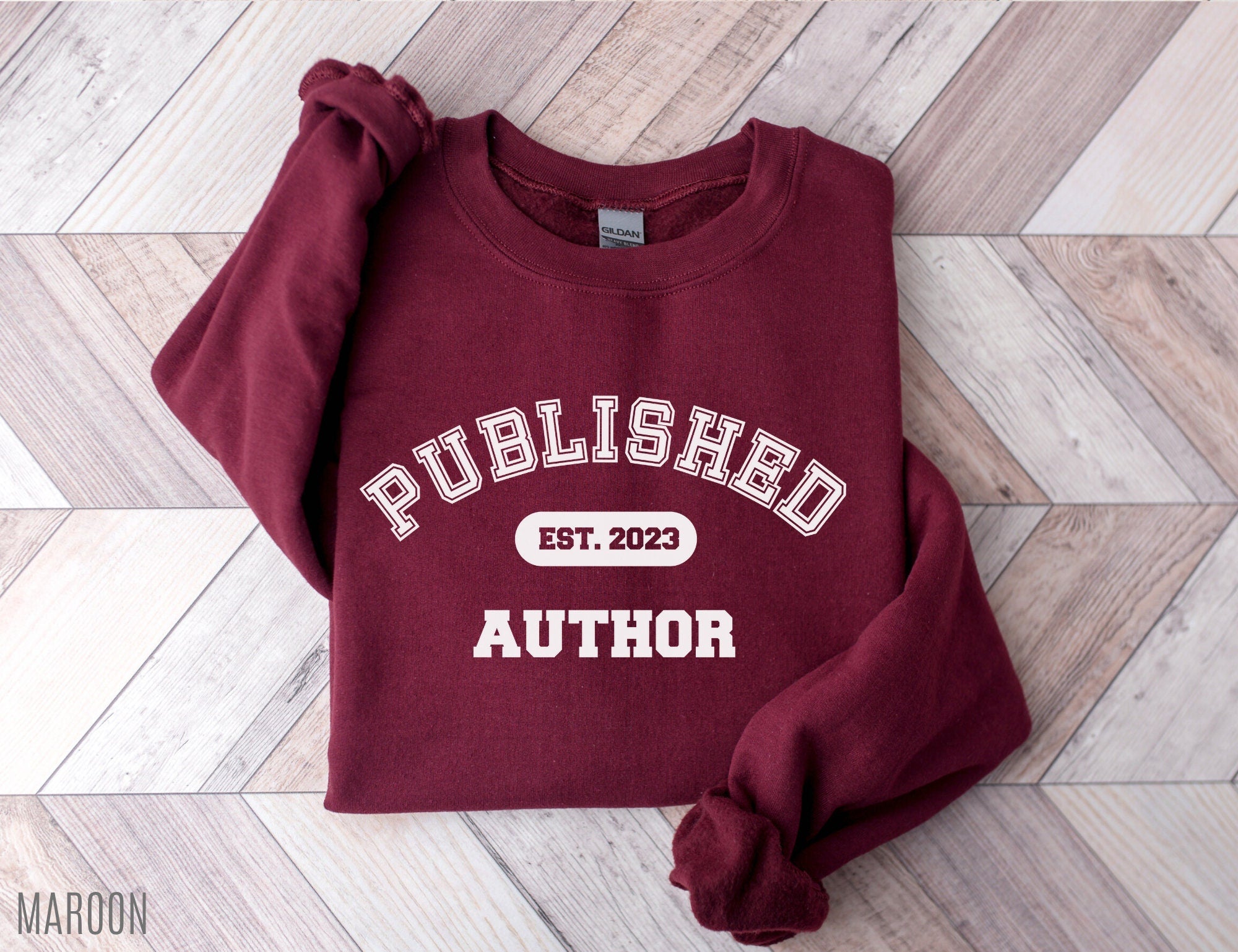 folded maroon sweatshirt that says published author est 2023 on chevron wood floor