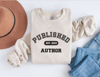 folded sand sweatshirt that says published author est 2023 styled with jeans and brown hat